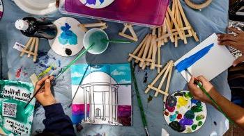 Overhead image with two people's hands seen finishing paintings, one of which is of the Old Well.